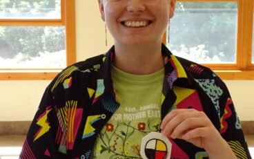 Picture of non-binary person with bright yellow-green hair and loud shirt holding a patch