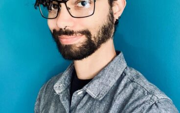 A professional headshot of CJ Harris, a person with dark curly hair and glasses, wearing a grey button-down shirt and smiling in front of a bright blue background.